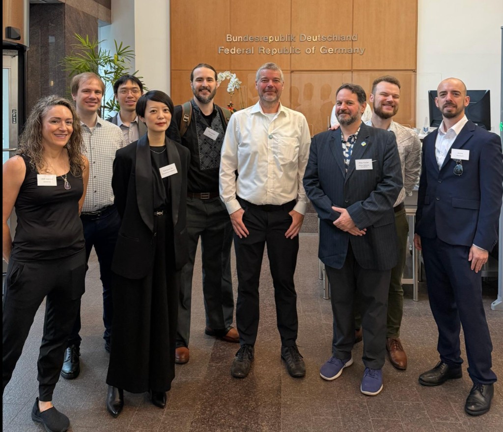 A group of open source maintainers and delegates pose together in the lobby of the German house in New York City, with Bundesrepublik Deutschland signage on the wall behind them. Pictured among the group are Jaime from conda-forge, Mike Fiedler from PyPI, Amanda Casari from Google, Seth Larson from PSF Security, and Leah Wasser.